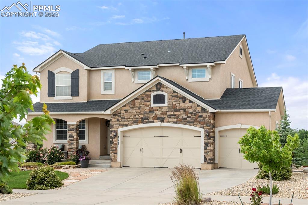 Caption: View of front of home with stone siding, a porch, stucco siding, concrete driveway, and a shingled r