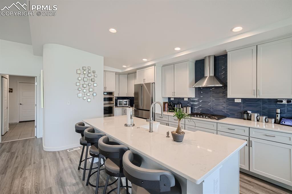 Image 6 of 31: Kitchen with backsplash, wall chimney range hood, light stone counters, sta