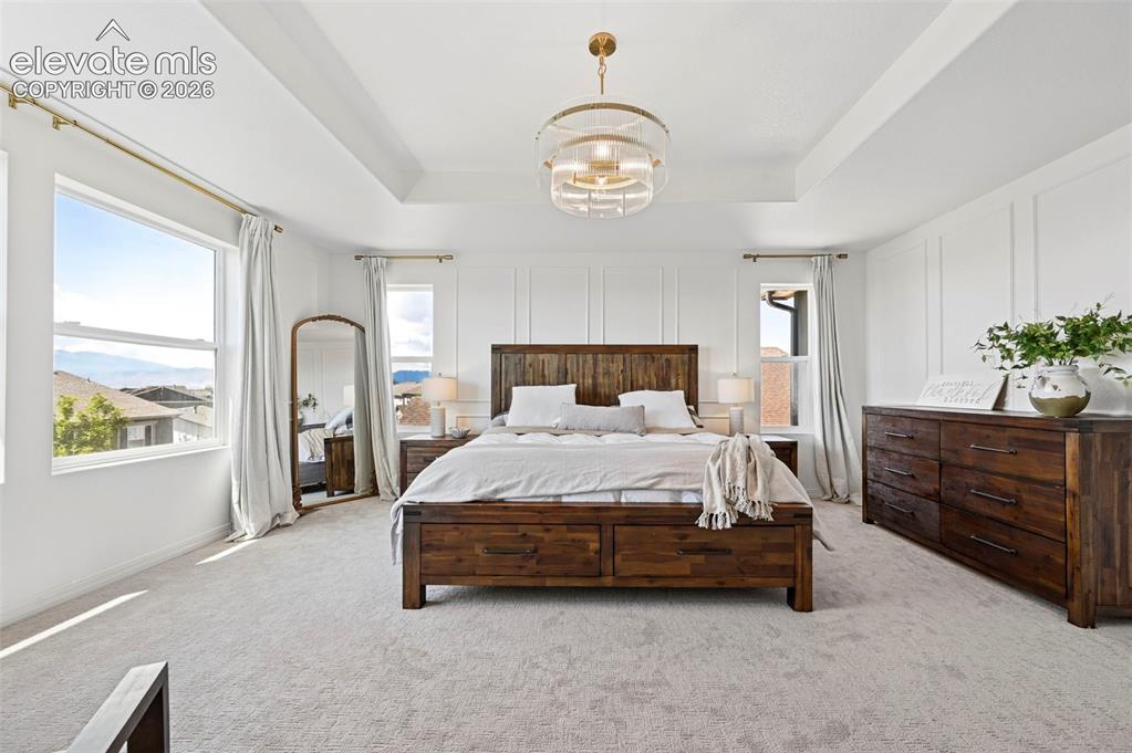 Image 45 of 50: Tray ceiling with a modern chandelier, wall paneling, and large windows