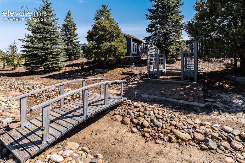 Image 47 of 50: Backyard bridge, arched trellis, and garden shed behind the trees.