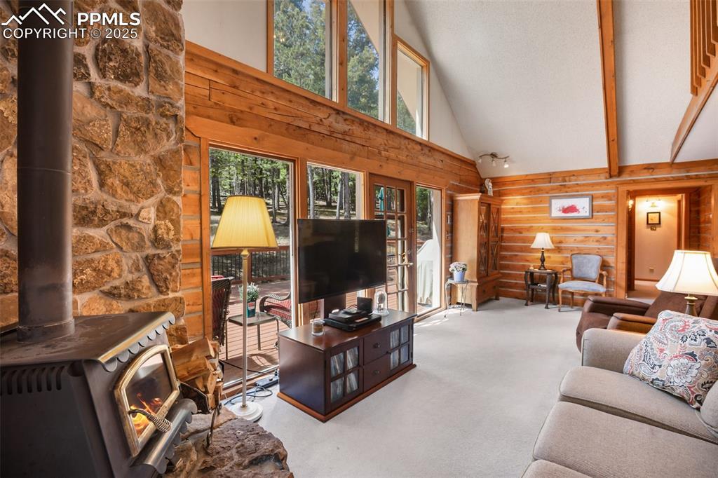 Image 11 of 36: Living area with high vaulted ceiling, carpet, a wood stove, wood walls, an