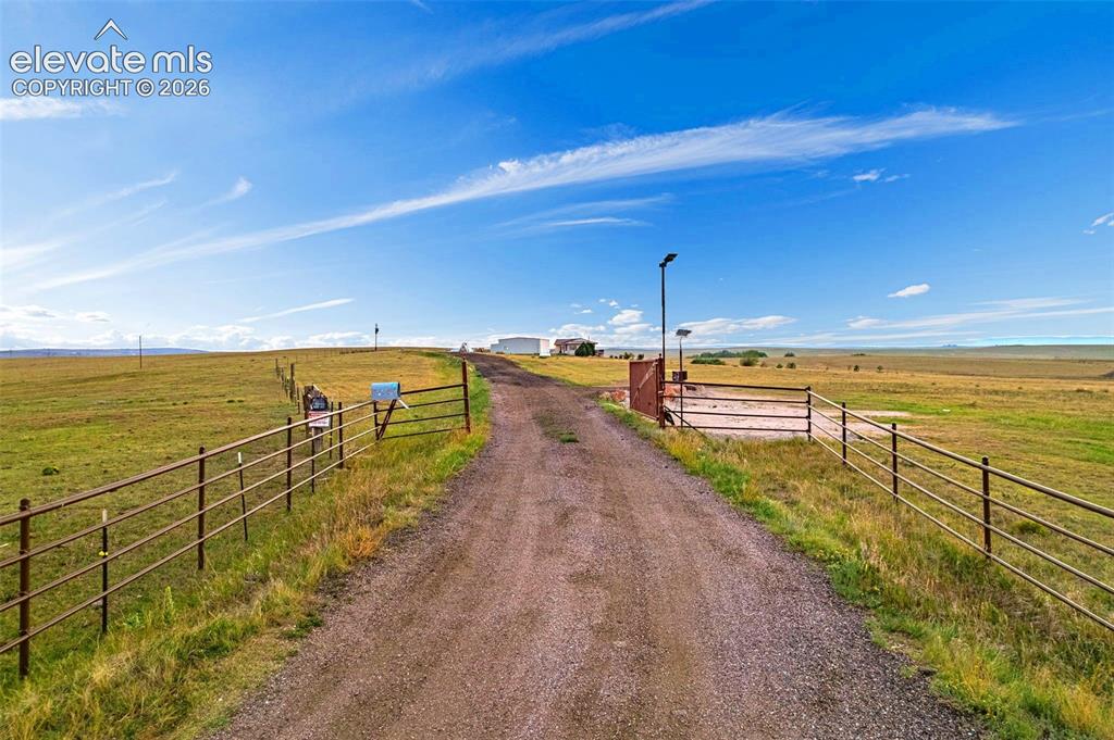 Image 6 of 50: Expansive open land with a gravel driveway, metal fencing, and a main gate