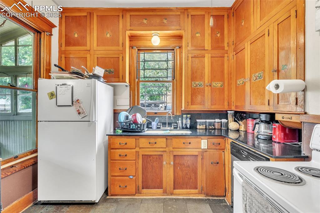 Image 15 of 49: Kitchen with white appliances, brown cabinets, dark countertops, and backsp