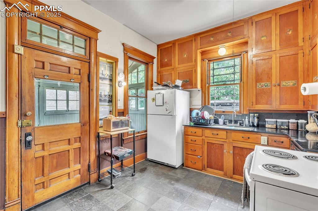 Image 16 of 49: Kitchen with white appliances, brown cabinetry, and dark countertops