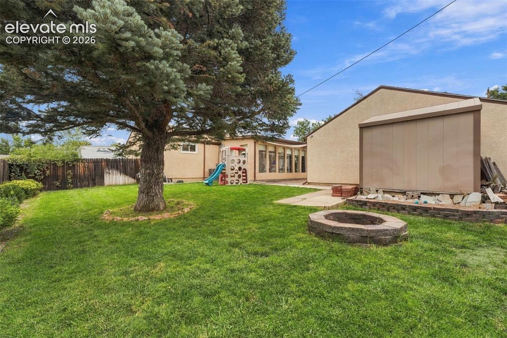Image 44 of 48: Expansive lawn with a mature tree, stone-built fire pit, and partial fencin