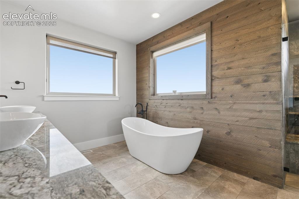 Image 26 of 50: Freestanding soaking tub, wood-finish accent wall, dual vessel sinks, stone