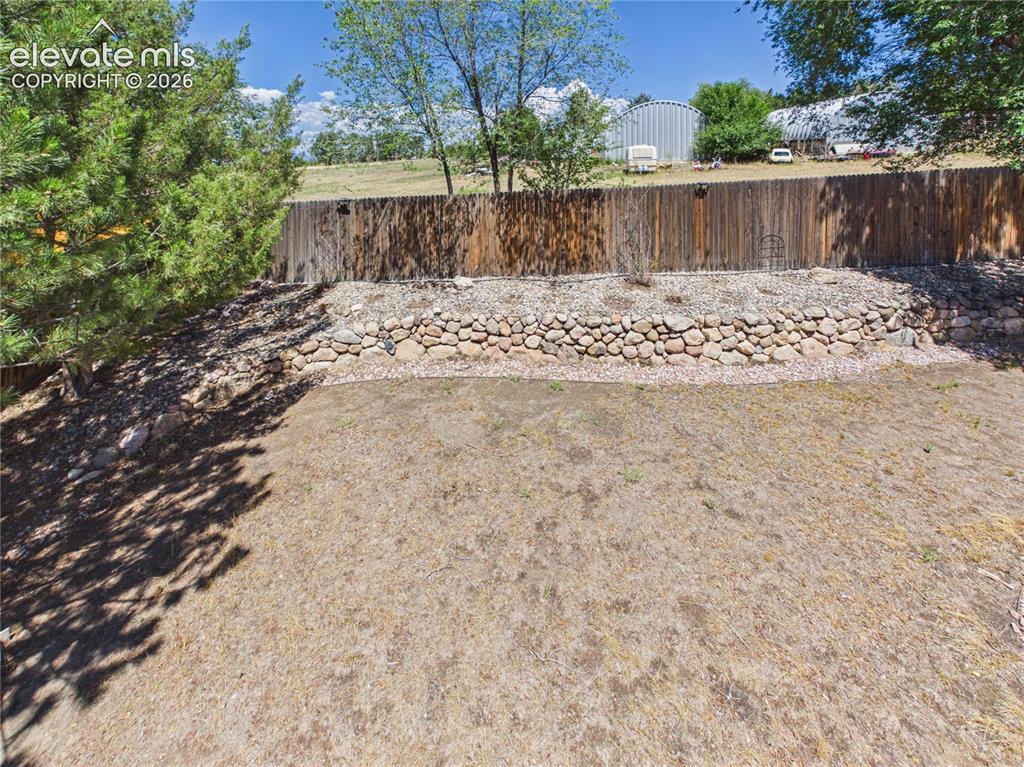 Image 36 of 46: Spacious fenced backyard with mature trees, extensive rock landscaping, and