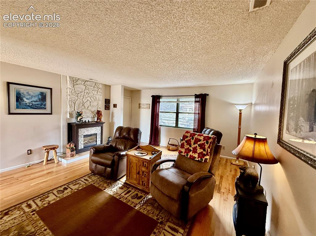 Image 8 of 26: Living area with wood finished floors, a textured ceiling, a stone fireplac