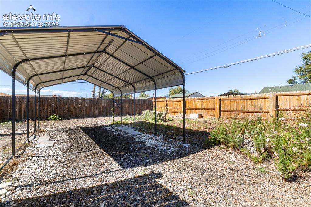 Image 22 of 23: Metal-frame carport with a corrugated roof, gravel ground cover, and a wood