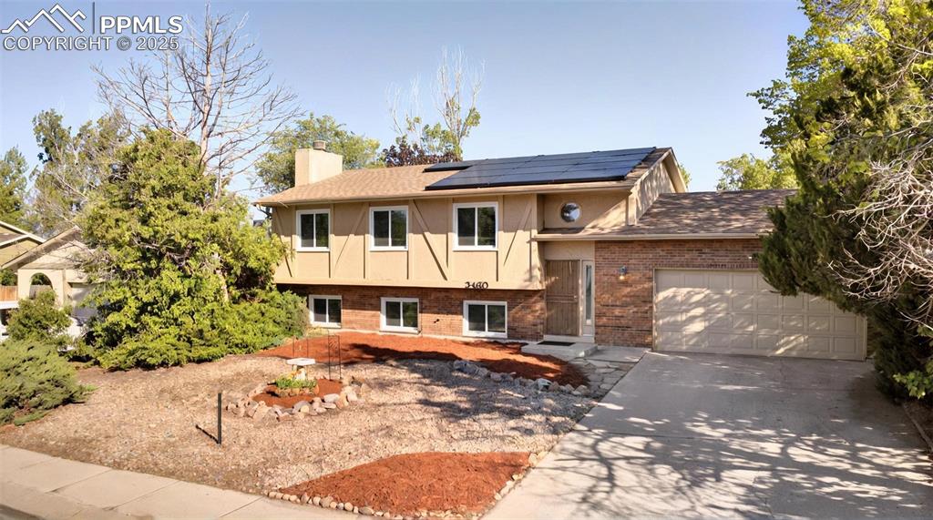 Caption: Raised ranch featuring brick siding, a chimney, solar panels, concrete driveway, and an attached gar