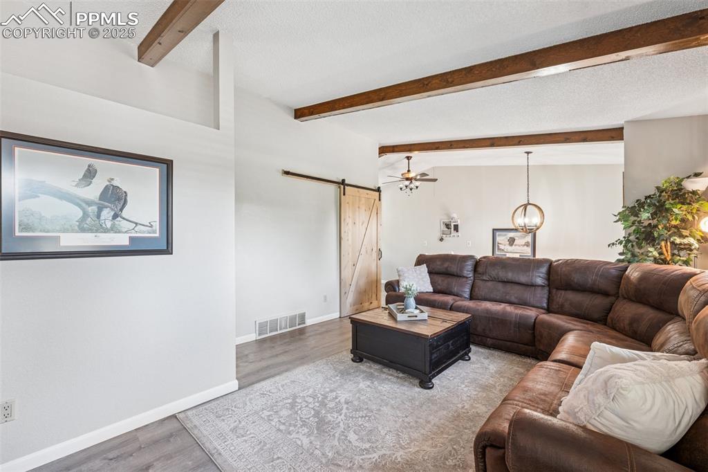 Image 9 of 25: Living room with a barn door, beamed ceiling, wood finished floors, and cei