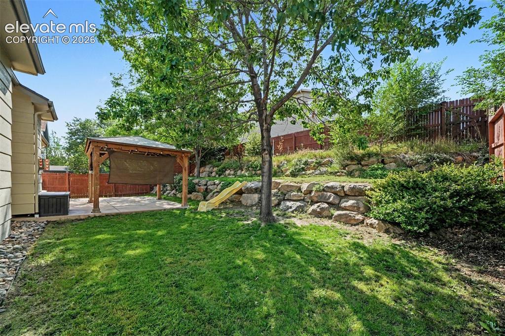 Image 26 of 28: The backyard features lush green grass, a fun slide, and mature tree provid