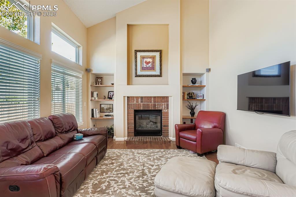Image 11 of 32: Living area with wood finished floors, high vaulted ceiling, a brick firepl