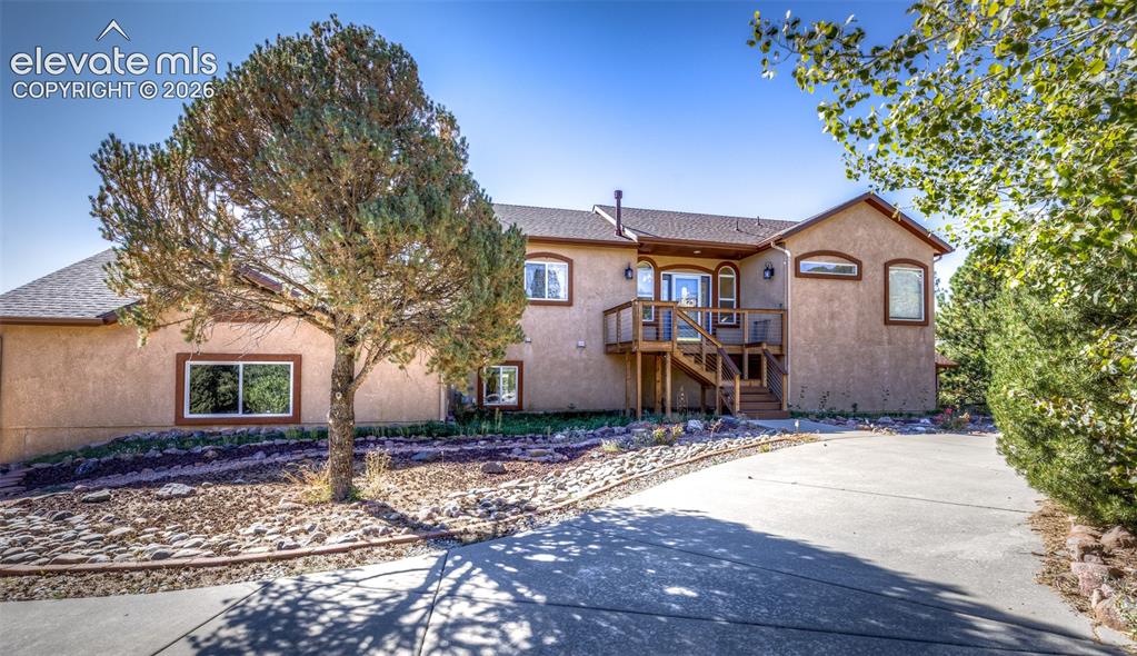 Caption: Stucco exterior featuring an elevated wood deck, concrete driveway, stone-accented landscape beds