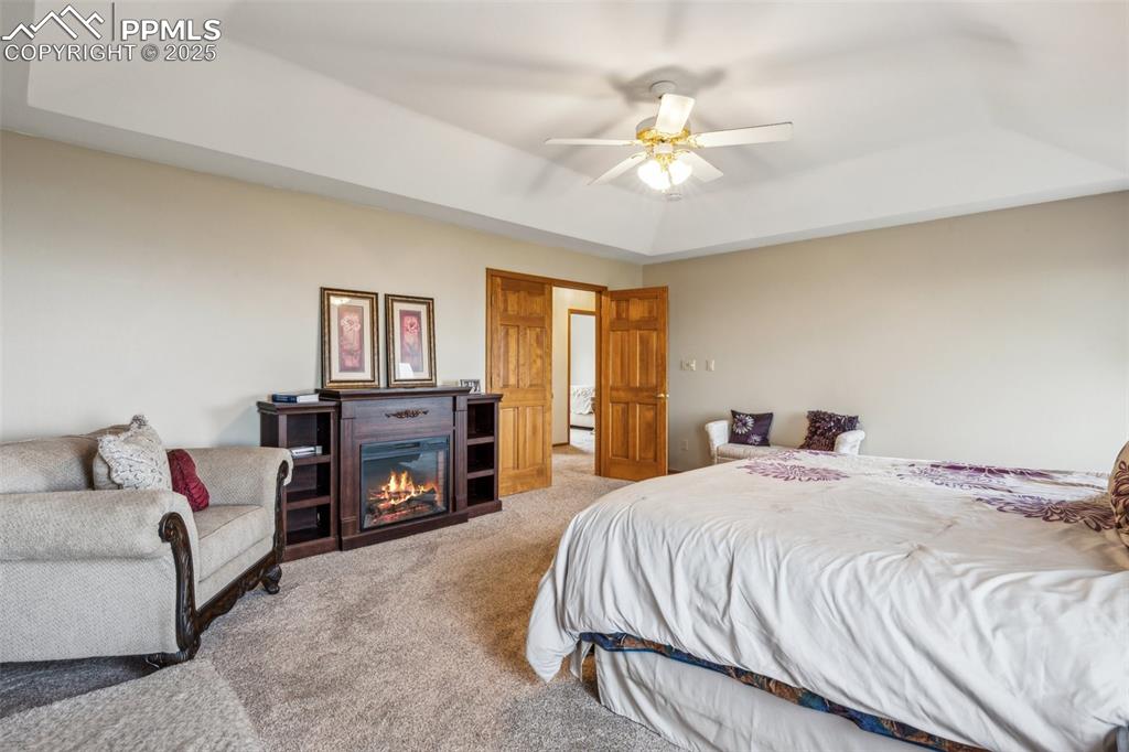 Image 26 of 43: Carpeted bedroom with a tray ceiling, ceiling fan, and a glass covered fire
