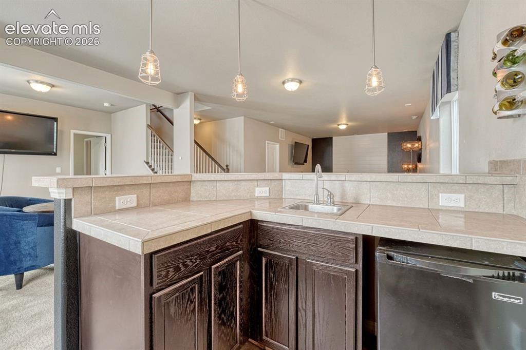 Image 36 of 50: Wet bar featuring dark wood-finish cabinetry, a tile countertop, and a stai