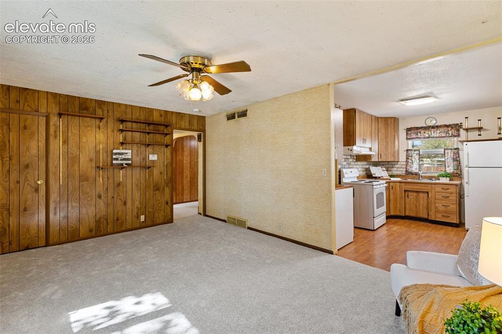 Image 11 of 45: Wood paneling accents a living area with a ceiling fan, opening to a kitche
