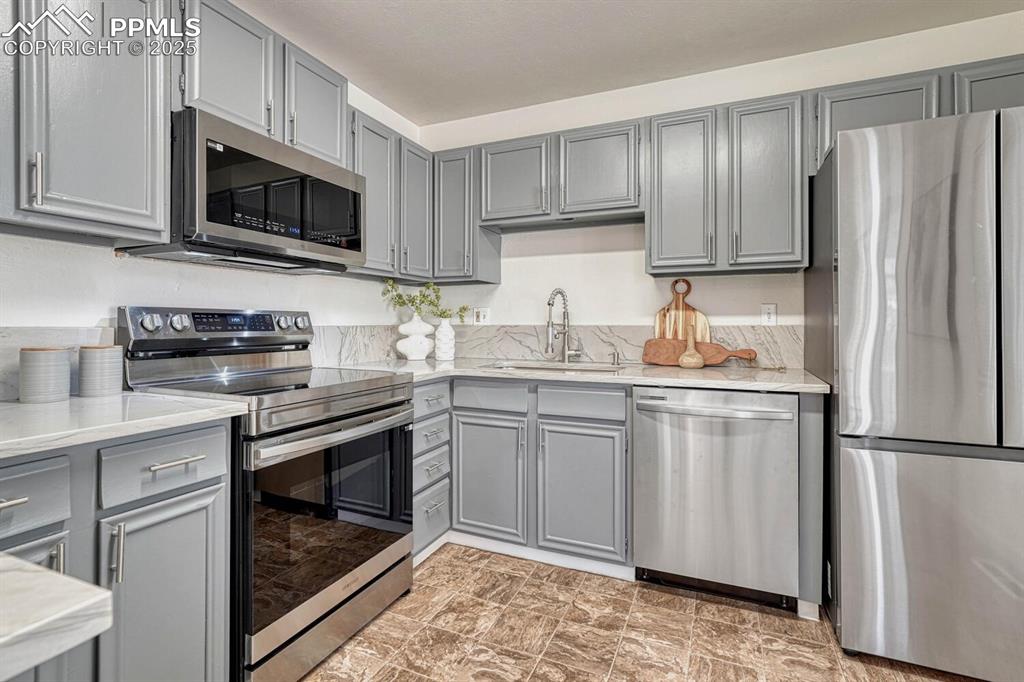 Image 12 of 43: Kitchen featuring gray cabinets, stainless steel appliances, and light ston