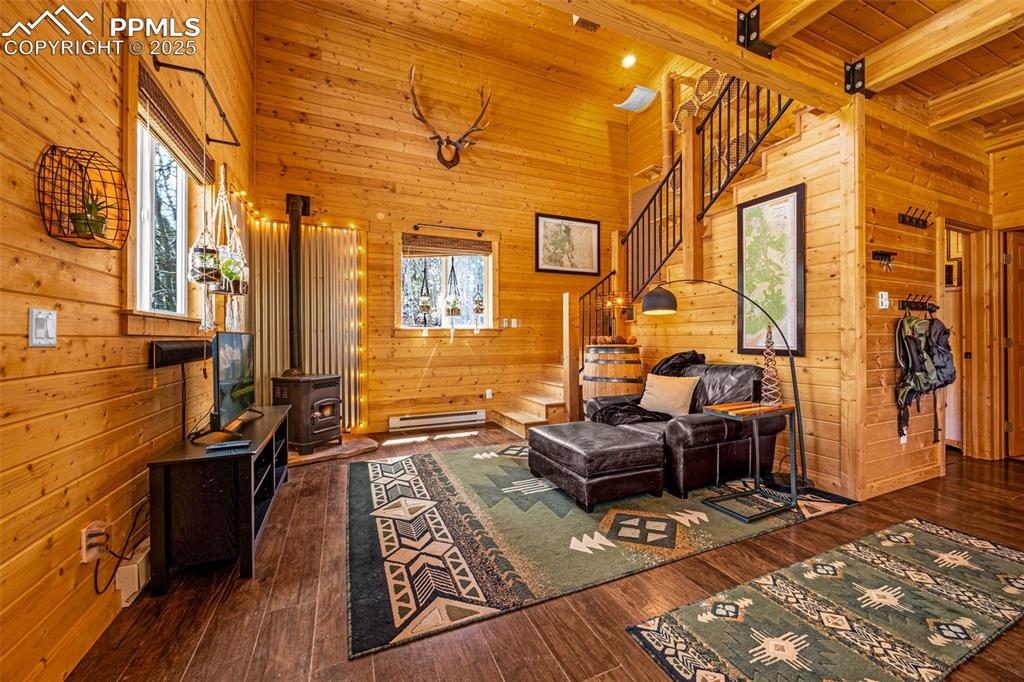 Image 6 of 45: Living room with baseboard heating, wood walls, a wood stove, stairway, and