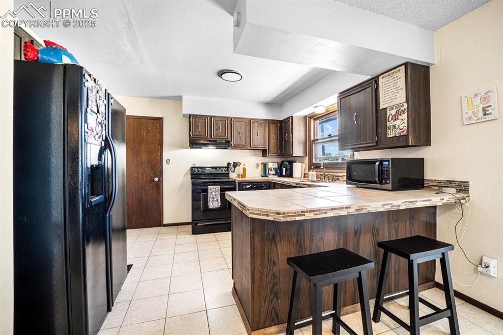 Image 8 of 38: Kitchen with dark brown cabinetry, tile counters, black appliances, light t