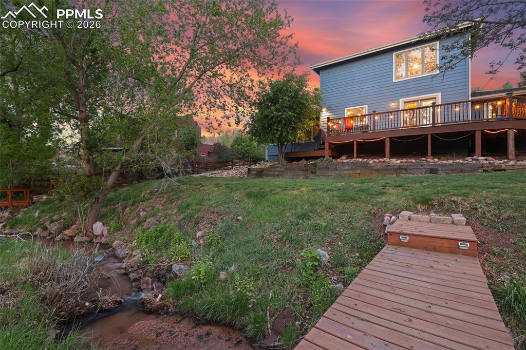 Image 9 of 47: Twilight photos of back of structure featuring bridge over backyard creek