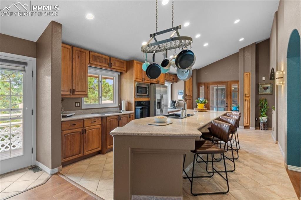 Image 11 of 49: Kitchen featuring a sink, black appliances, an island with sink, brown cabi
