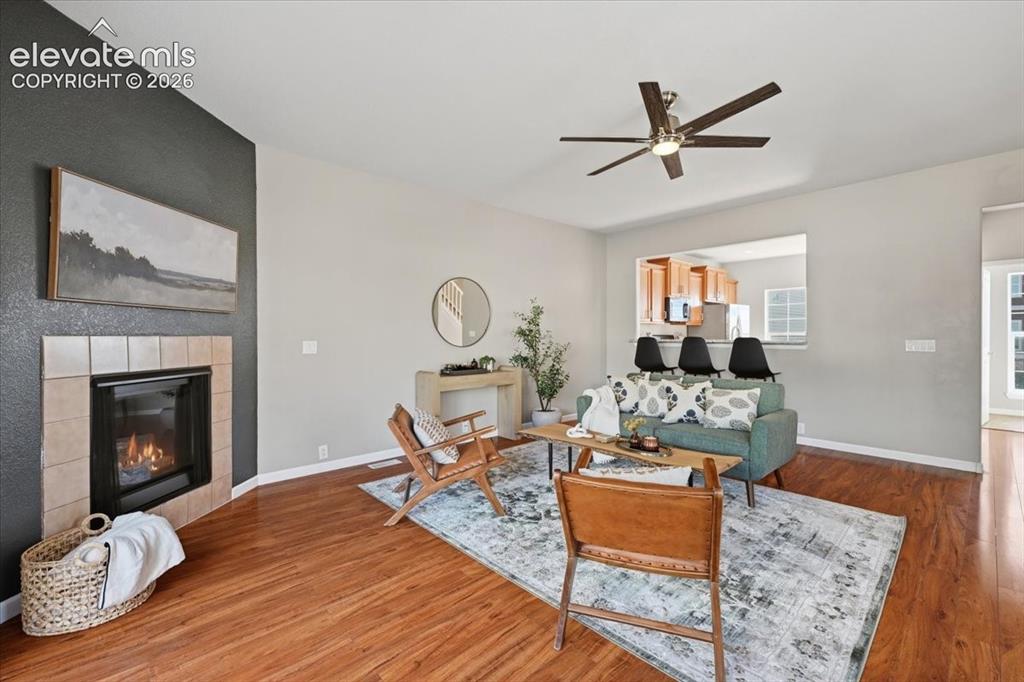 Image 10 of 34: Hardwood flooring, tiled fireplace, ceiling fan, and a kitchen pass-through