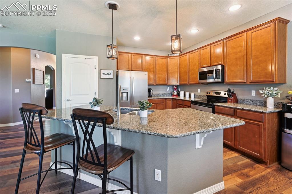 Image 4 of 39: Kitchen with arched walkways, dark wood-style flooring, brown cabinetry, ap