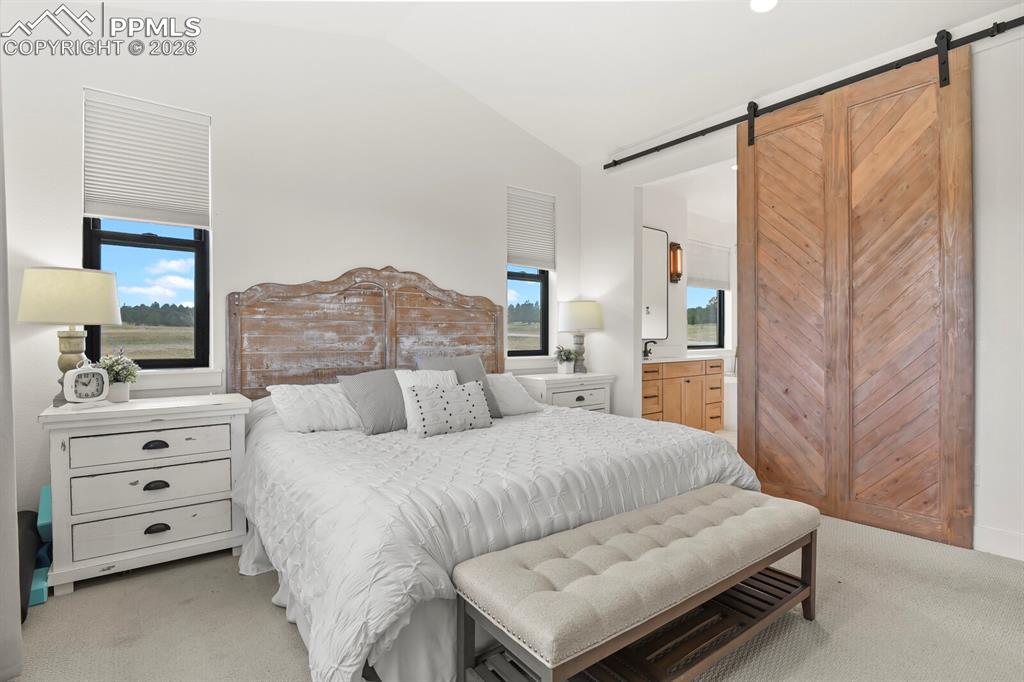 Image 21 of 47: Bedroom featuring a barn door, vaulted ceiling, light carpet, and ensuite b