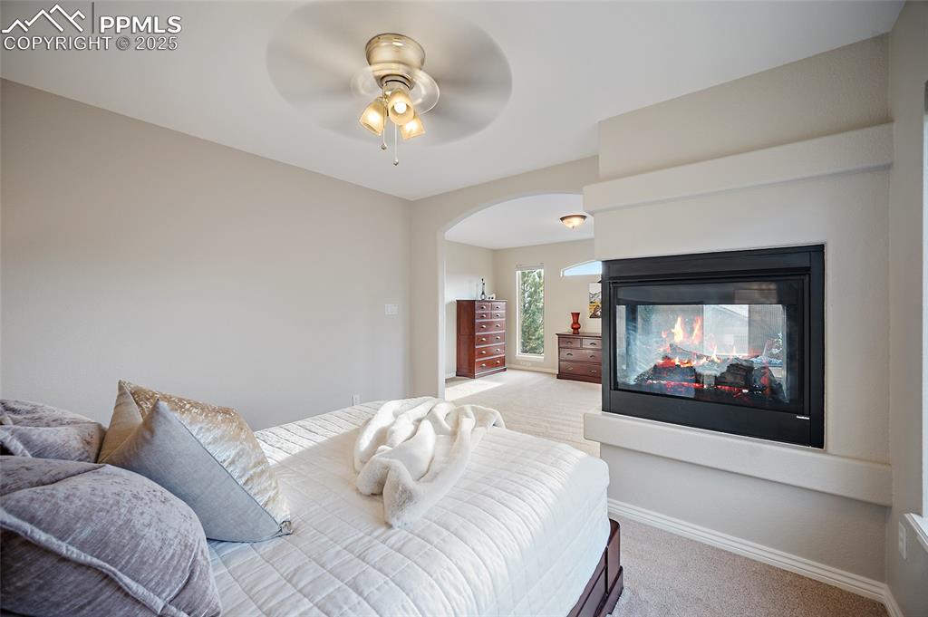 Image 24 of 50: Bedroom with light colored carpet, a multi sided fireplace, a ceiling fan, 