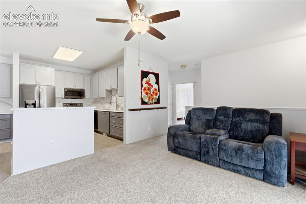 Image 10 of 25: Open-concept living space with light-toned carpeting and a ceiling fan