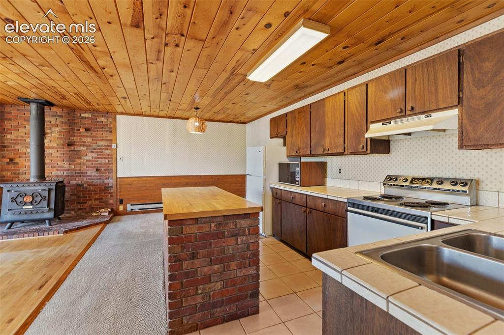 Image 13 of 46: Kitchen and living area featuring a wood-panel ceiling, brick wall with a w