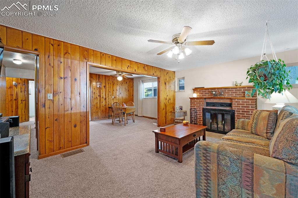 Image 6 of 27: Carpeted living area featuring a brick fireplace, wood walls, a textured ce