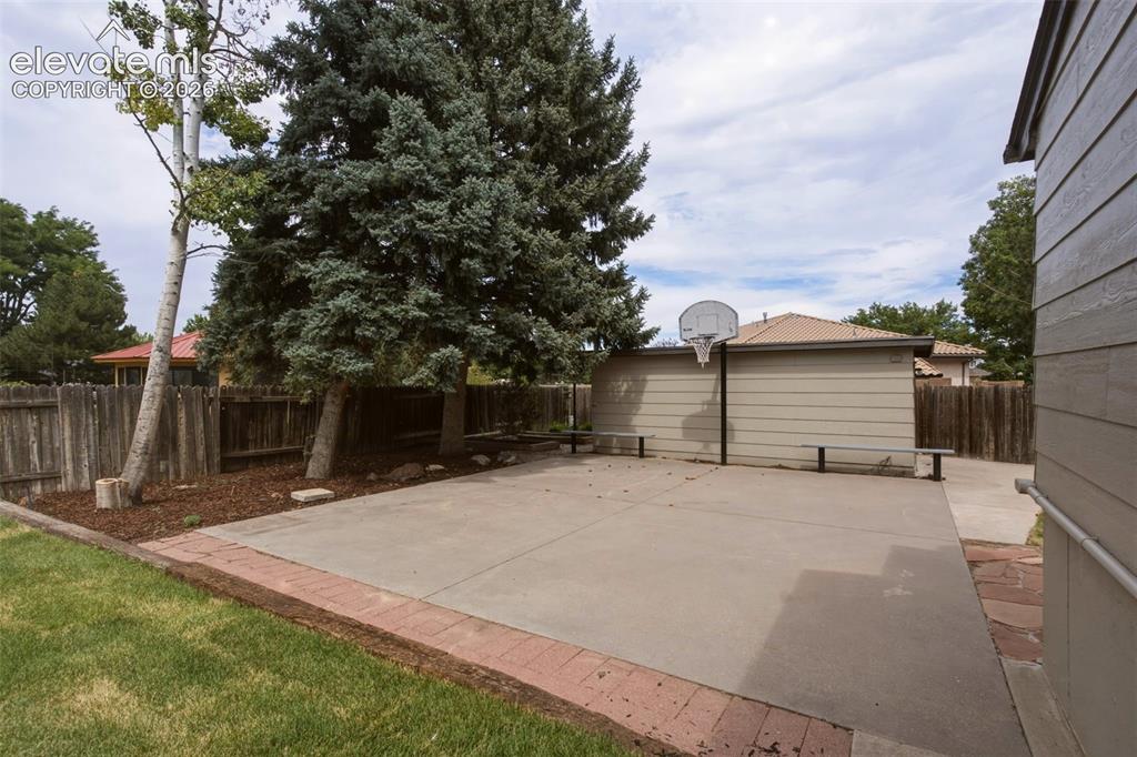 Image 29 of 32: Paved patio area featuring a basketball hoop, mature trees, and a privacy f