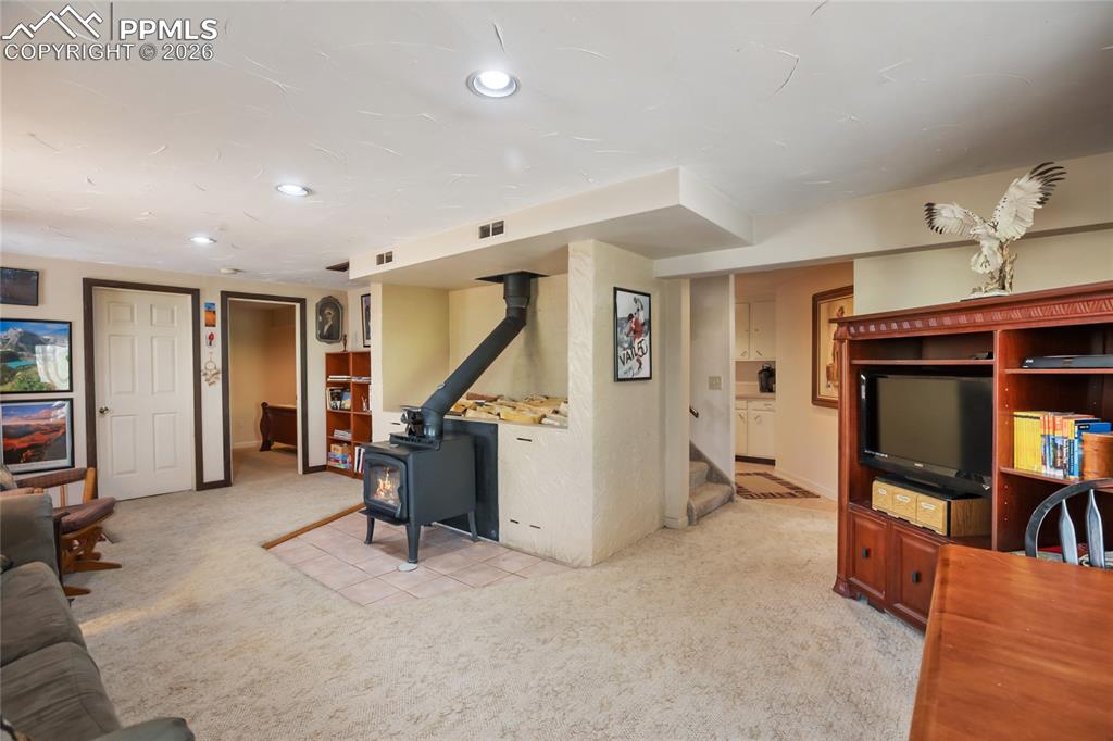 Image 18 of 39: The downstairs family room with a wood stove, light carpet, stairs, and rec