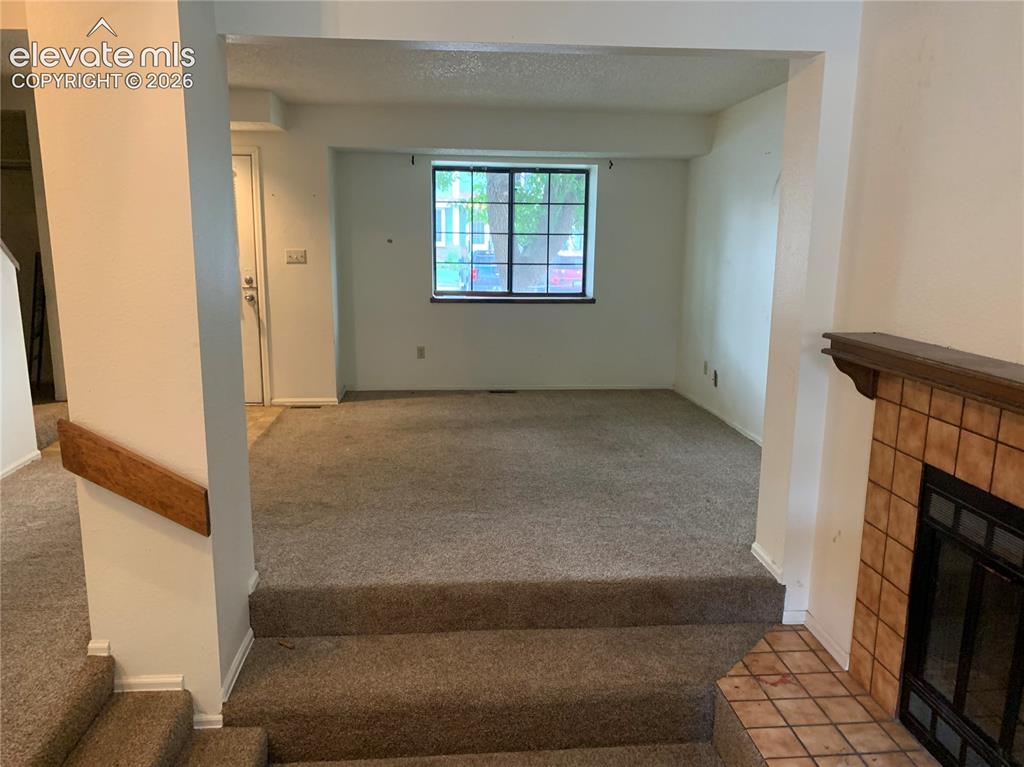 Image 17 of 27: Carpeted living area featuring a central window, white walls, and a tiled f