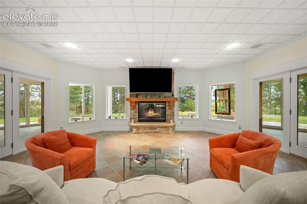 Image 28 of 50: Stone fireplace with wood mantel, large windows, recessed lighting, and til