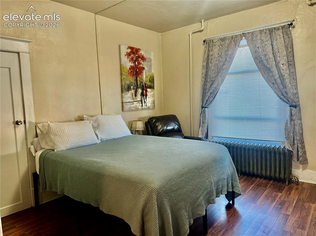 Image 5 of 50: Wood-finish flooring with a window featuring blinds and curtains, and a rad