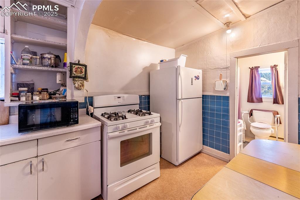 Image 8 of 21: Kitchen with white appliances, light countertops, tile walls, open shelves,