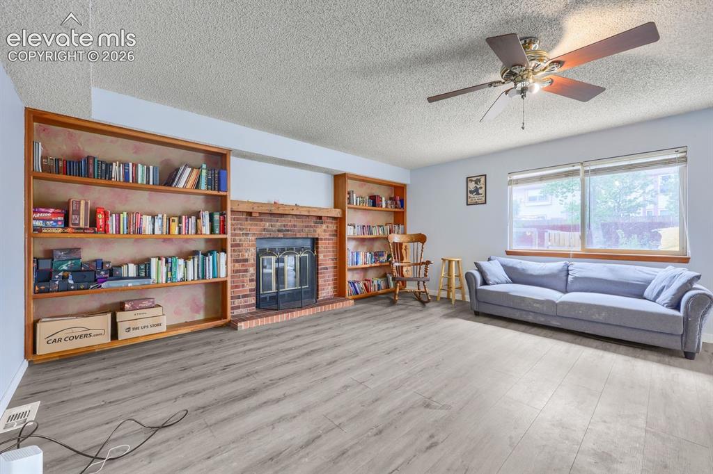 Image 10 of 42: Light wood-finish flooring, built-in shelving, brick fireplace, and ceiling
