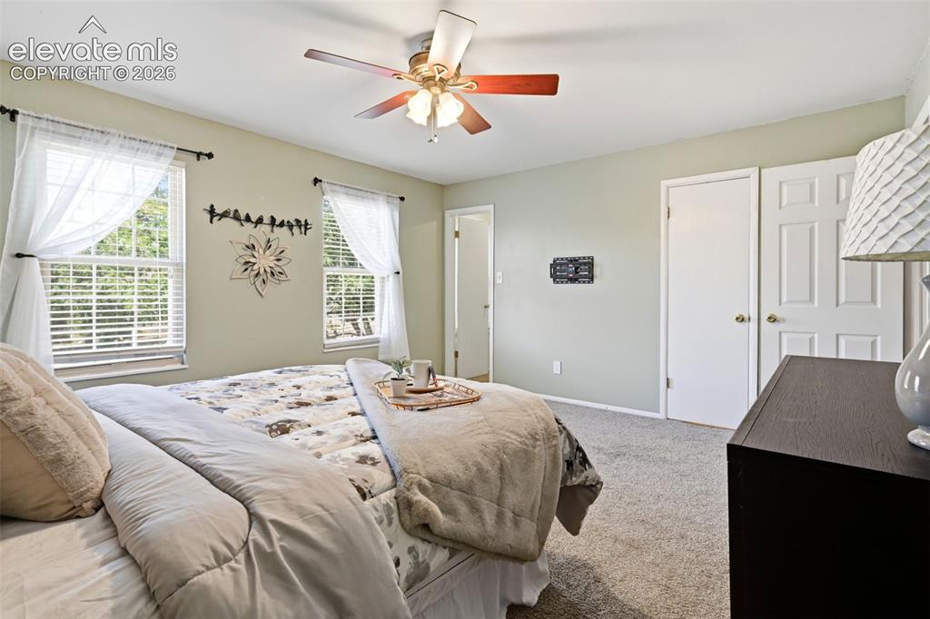 Image 32 of 50: Carpeted bedroom featuring two windows with blinds, a ceiling fan, and two