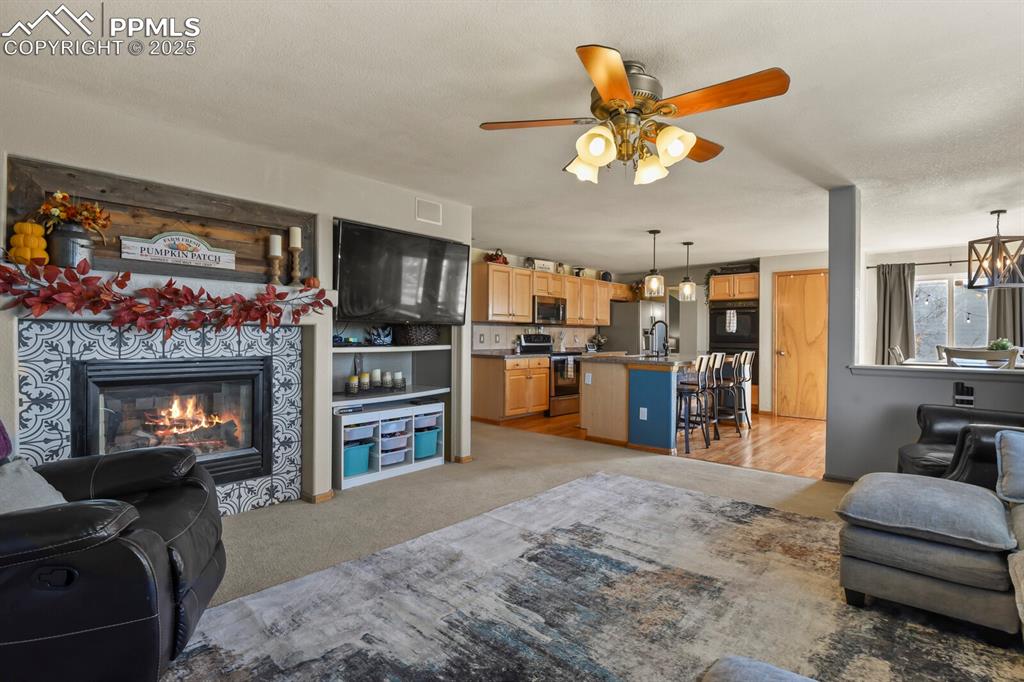 Image 18 of 50: Living area with light colored carpet, a tile fireplace, and ceiling fan