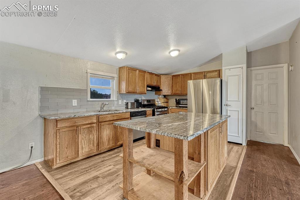 Image 8 of 38: Kitchen with a textured wall, tasteful backsplash, brown cabinetry, stainle