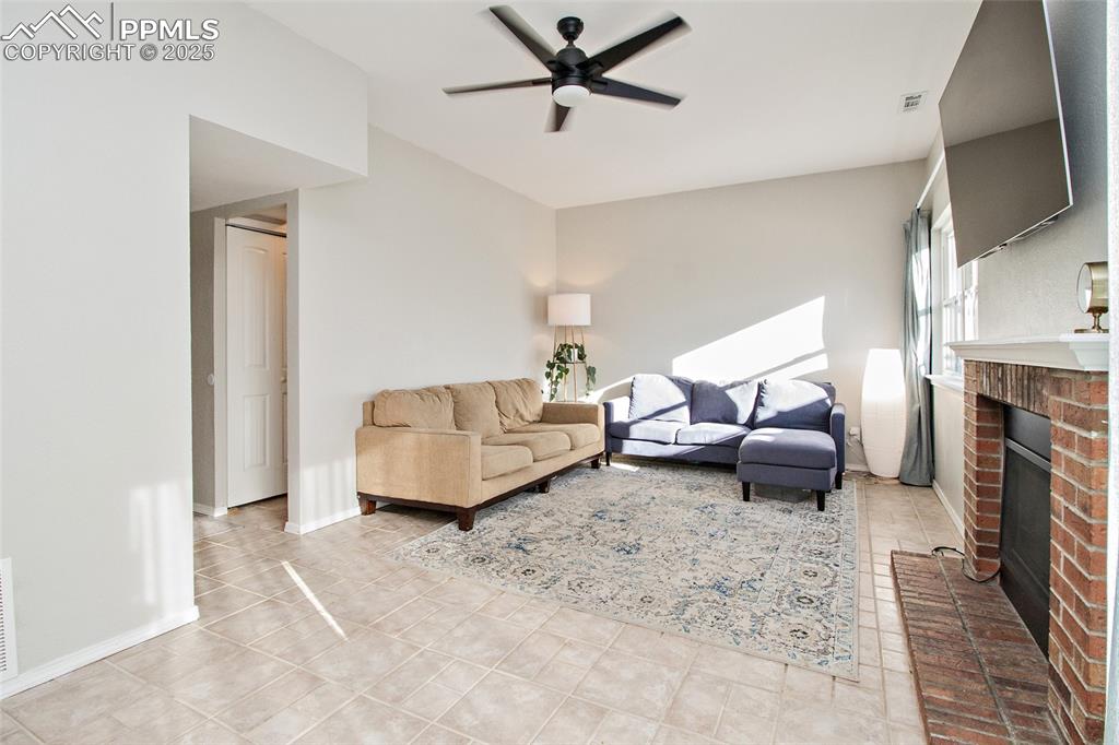 Image 25 of 43: Living room featuring a ceiling fan, a brick fireplace, and light tile patt