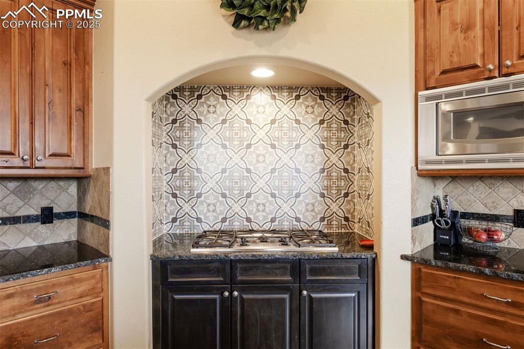 Image 6 of 50: Kitchen with decorative backsplash, dark stone counters, brown cabinetry, a