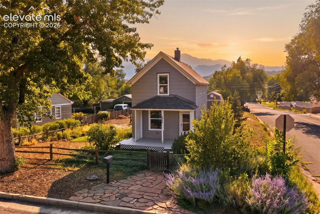 Caption: Gray siding exterior with white trim, covered front porch, stone pathway, mature trees, and mountain