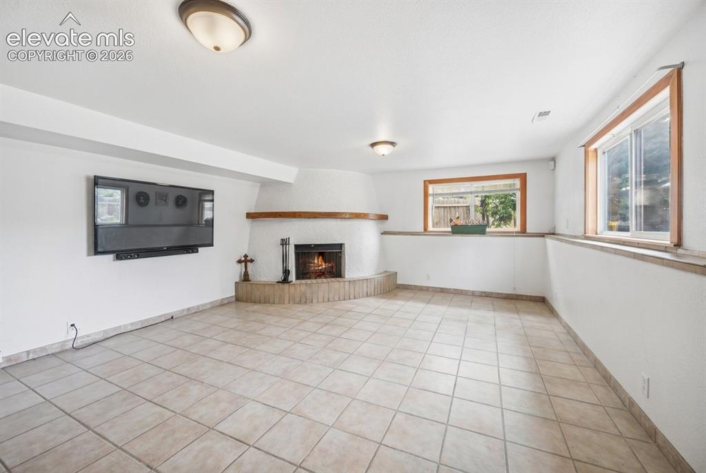 Image 13 of 20: Corner fireplace with a wood mantel, tile flooring, and multiple windows wi