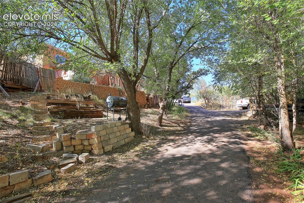 Image 20 of 26: Asphalt driveway surrounded by mature trees featuring segmented stone retai
