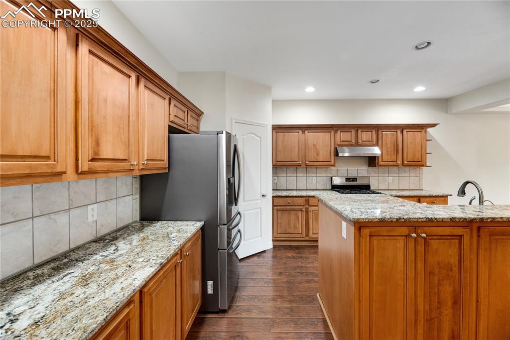 Image 39 of 48: Kitchen with light stone countertops, brown cabinetry, tasteful backsplash,