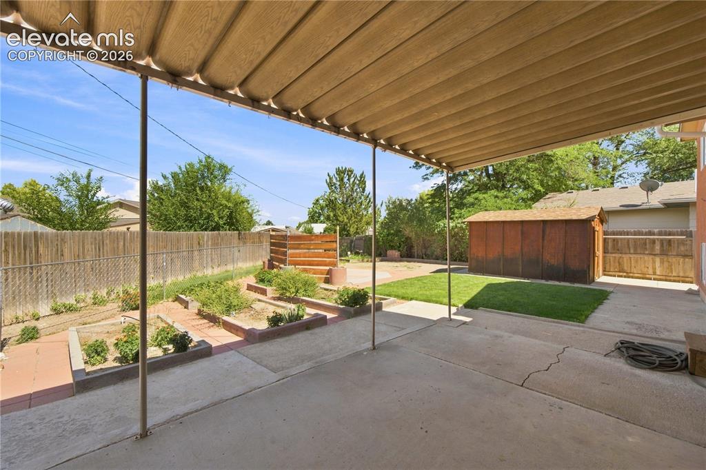 Image 25 of 39: Covered patio with concrete flooring, raised garden beds, and a storage she